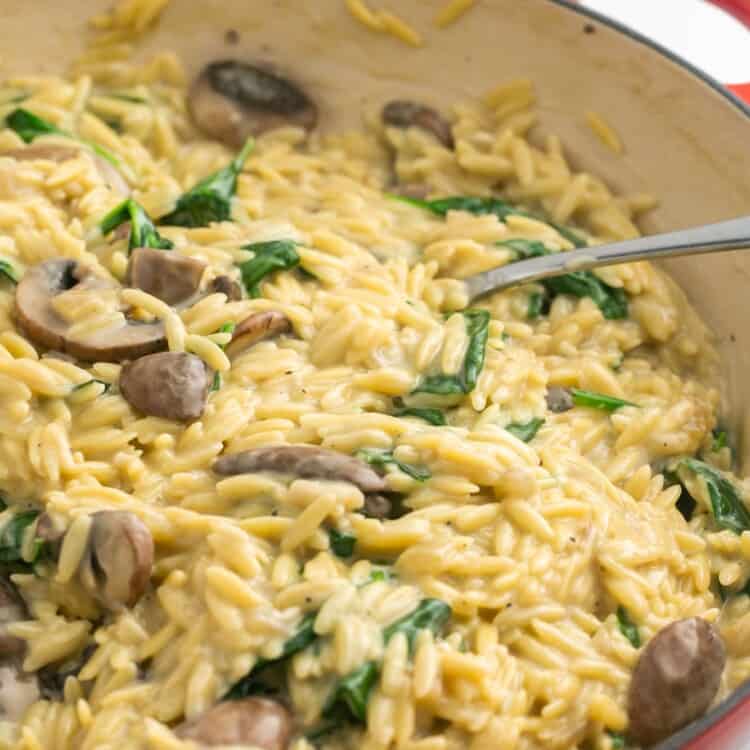 a red enameled skillet filled with creamy mushroom spinach orzo. A spoon is in the pan.