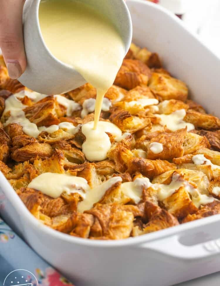 warm vanilla sauce being poured over croissant bread pudding in a casserole dish.