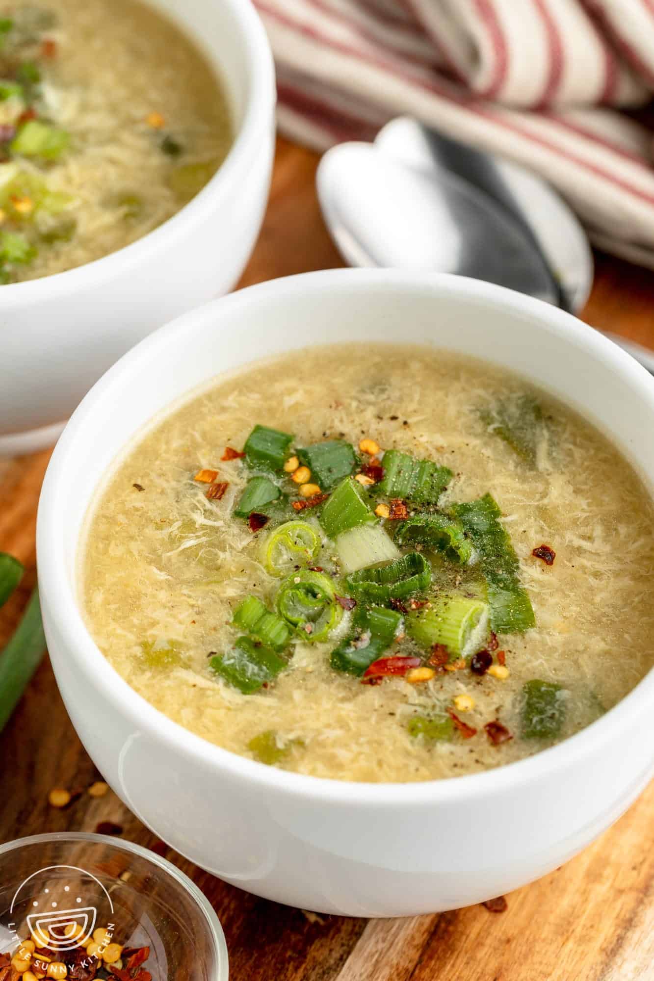 a bowl of homemade egg drop soup garnished with green onion, sitting on a wooden cutting board next to another bowl of soup and a small bowl of crushed red pepper flakes.