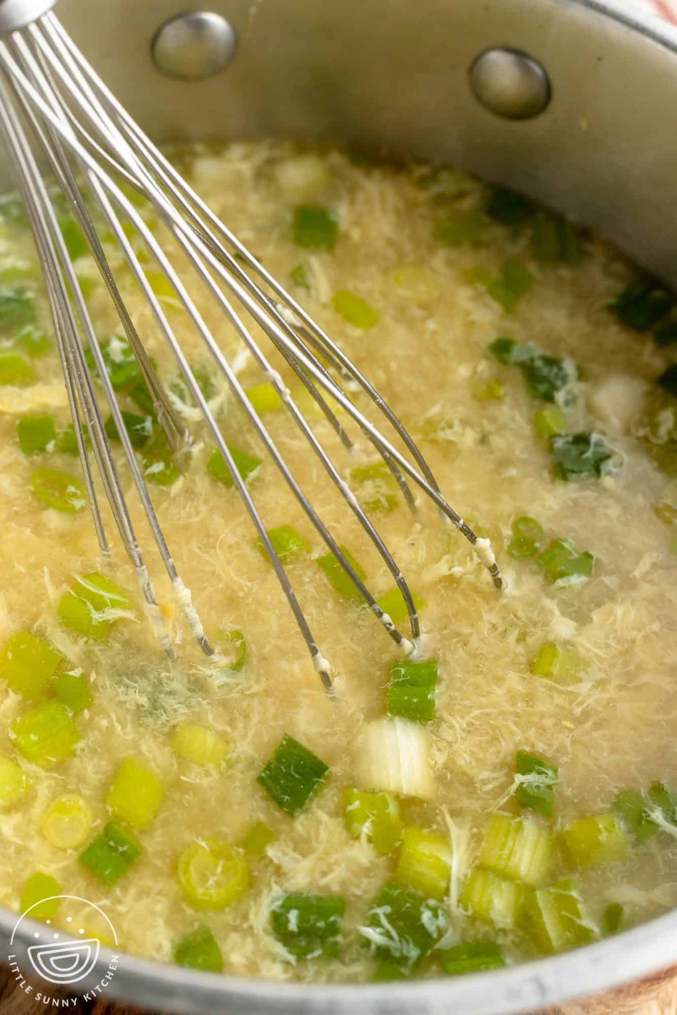 A pot of soup. A whisk is stirring in eggs to make egg drop soup ribbons.