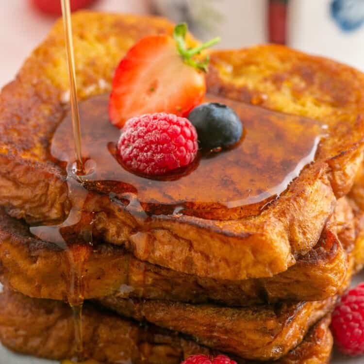 a plate of stacked slices of french toast with fresh berries and maple syrup on top.