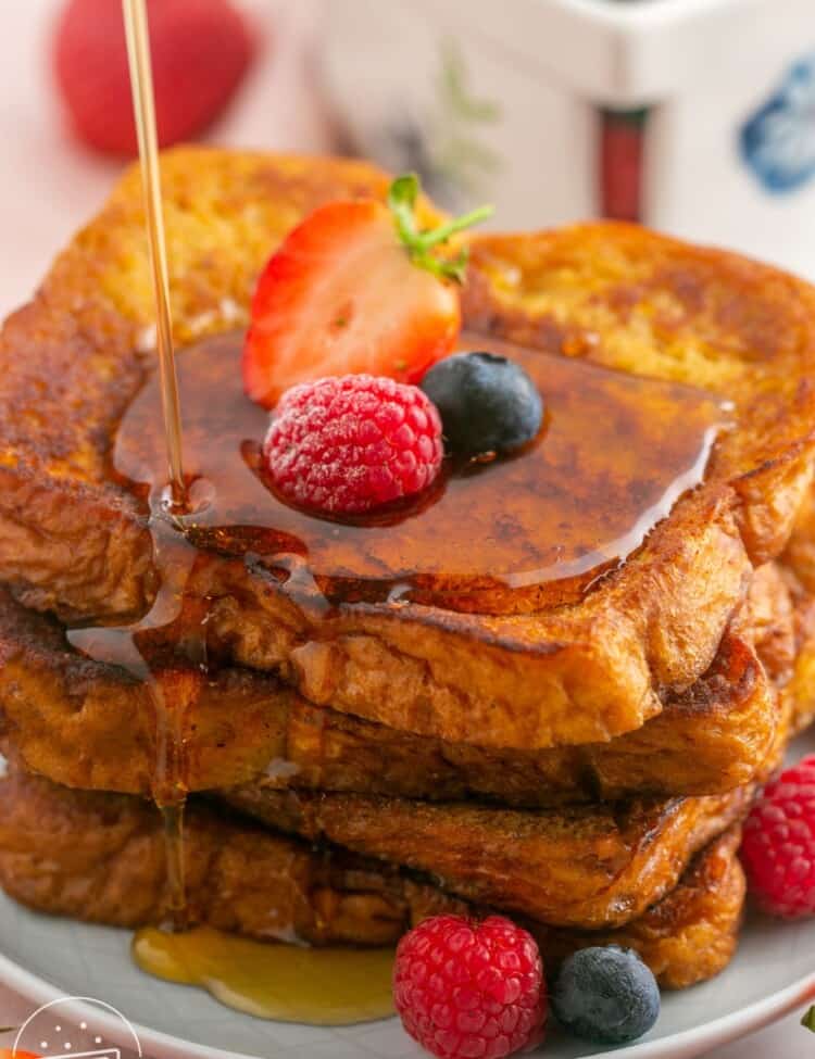 a plate of stacked slices of french toast with fresh berries and maple syrup on top.