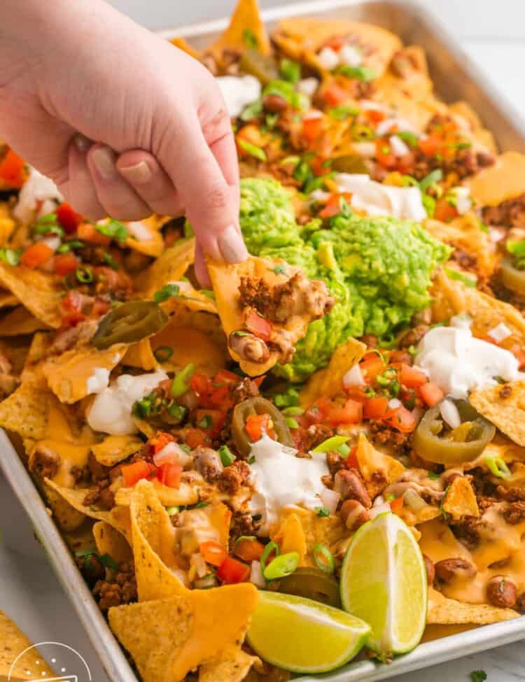 a tray of loaded nachos. A hand is picking up a chip topped with cheese and beef.