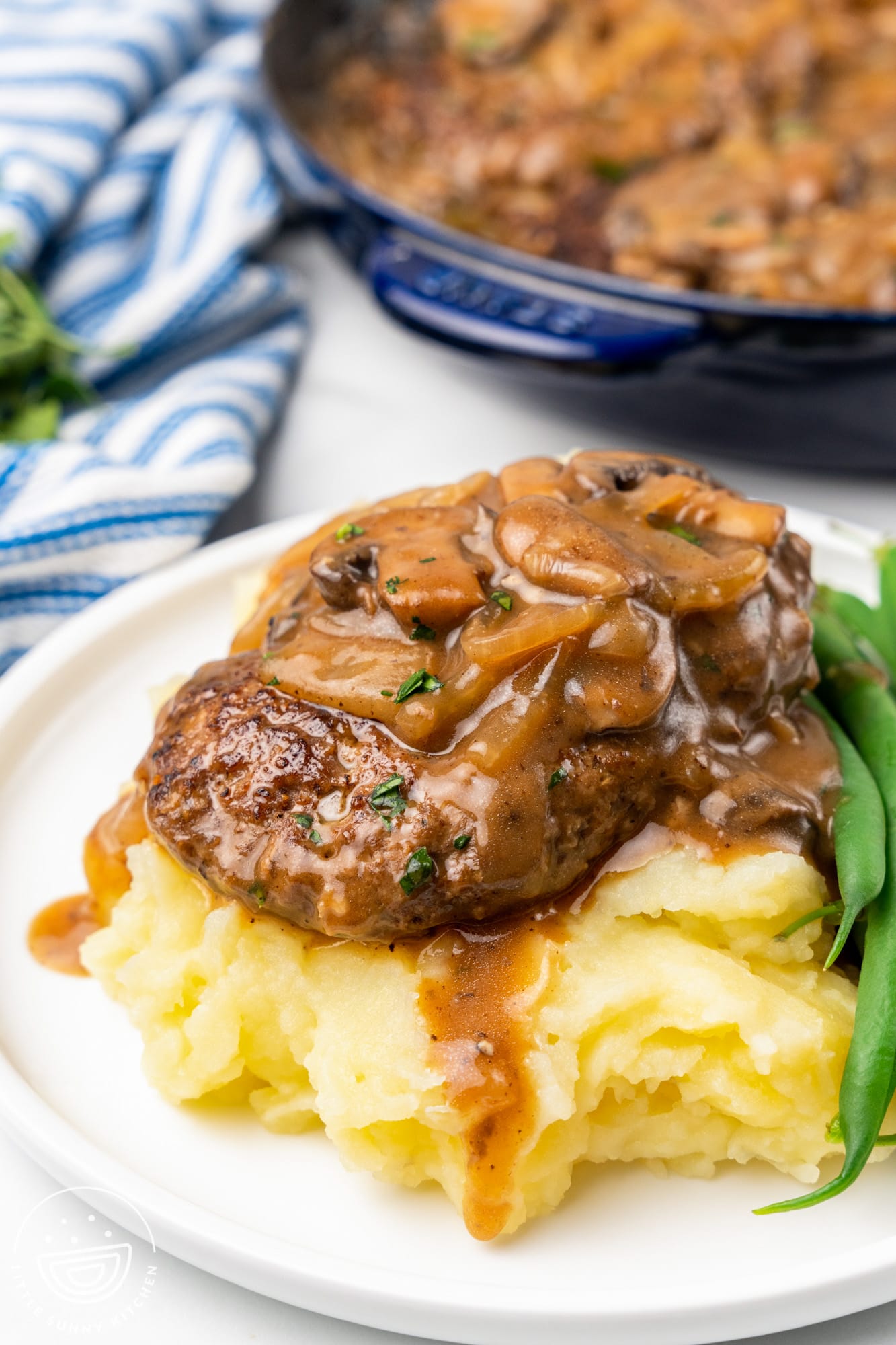 Hamburger steak sitting over mashed potatoes with mushroom gravy, and green beans on the side.