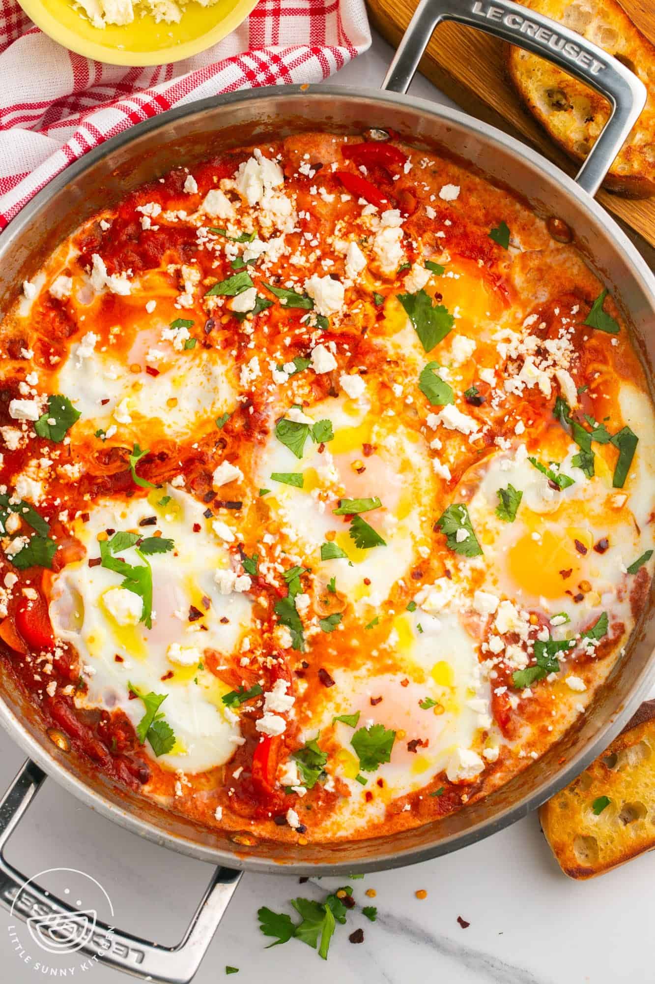 Overhead shot of shakshuka in a large stainless steel skillet, topped with crumbled feta cheese and chopped fresh parsley