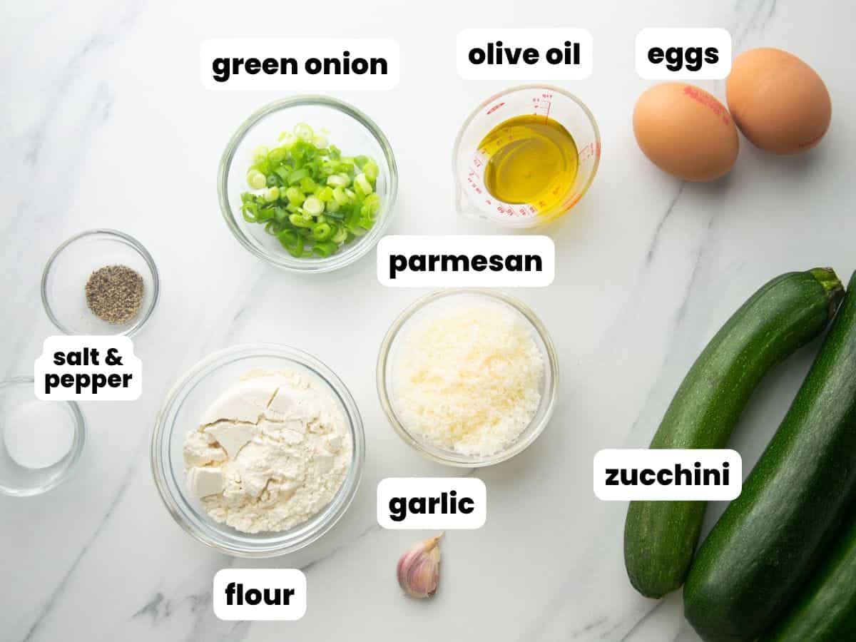 The ingredients for zucchini fritters, all in small bowls on a marble counter