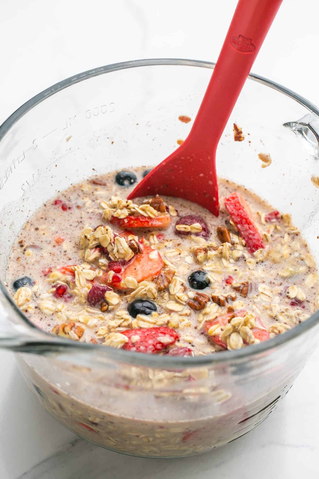 Oats, berries milk, and ingredients being mixed in a glass bowl to make baked oatmeal in a pan.