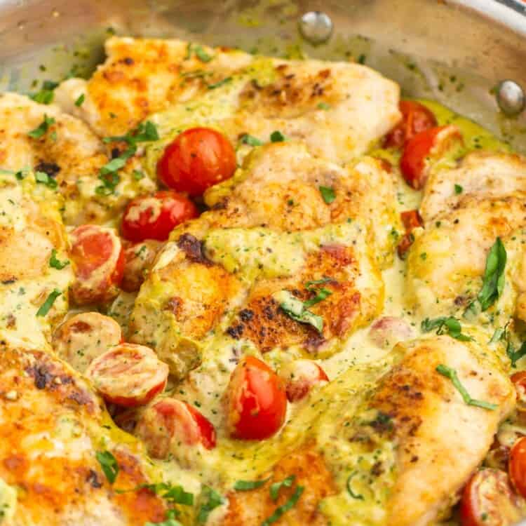 a stainless steel skillet filled with seared chicken breasts in a creamy pesto sauce, topped with halved cherry tomatoes.