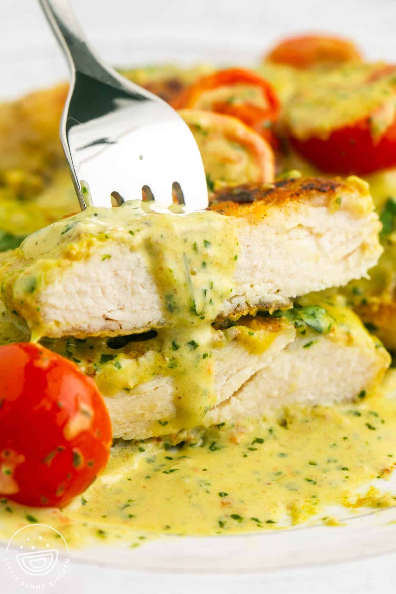 two slices of chicken held with a fork on a plate. The chicken is coated with a creamy pesto sauce.