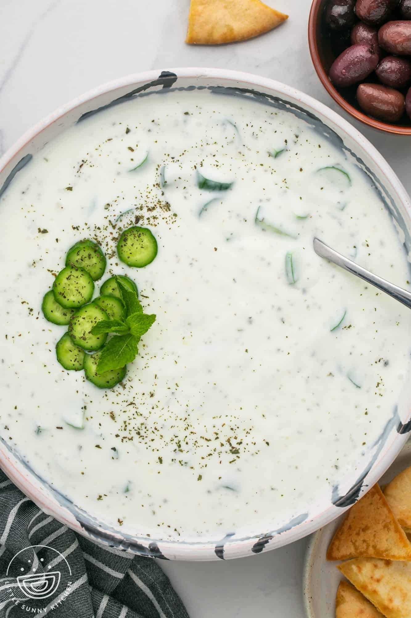 Overhead shot of khyar bi laban, or cucumber yogurt sauce in a large bowl, with kalmata olives on the side and pita chips.