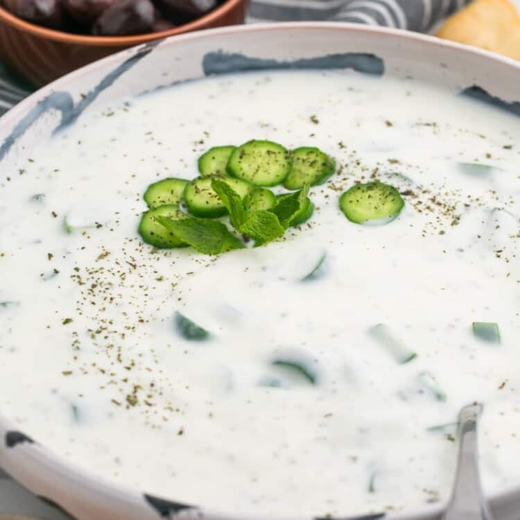 Cucumber yogurt sauce in a large bowl, with sliced of cucumber and dried mint, and a serving spoon. Kalamata olives in the background.
