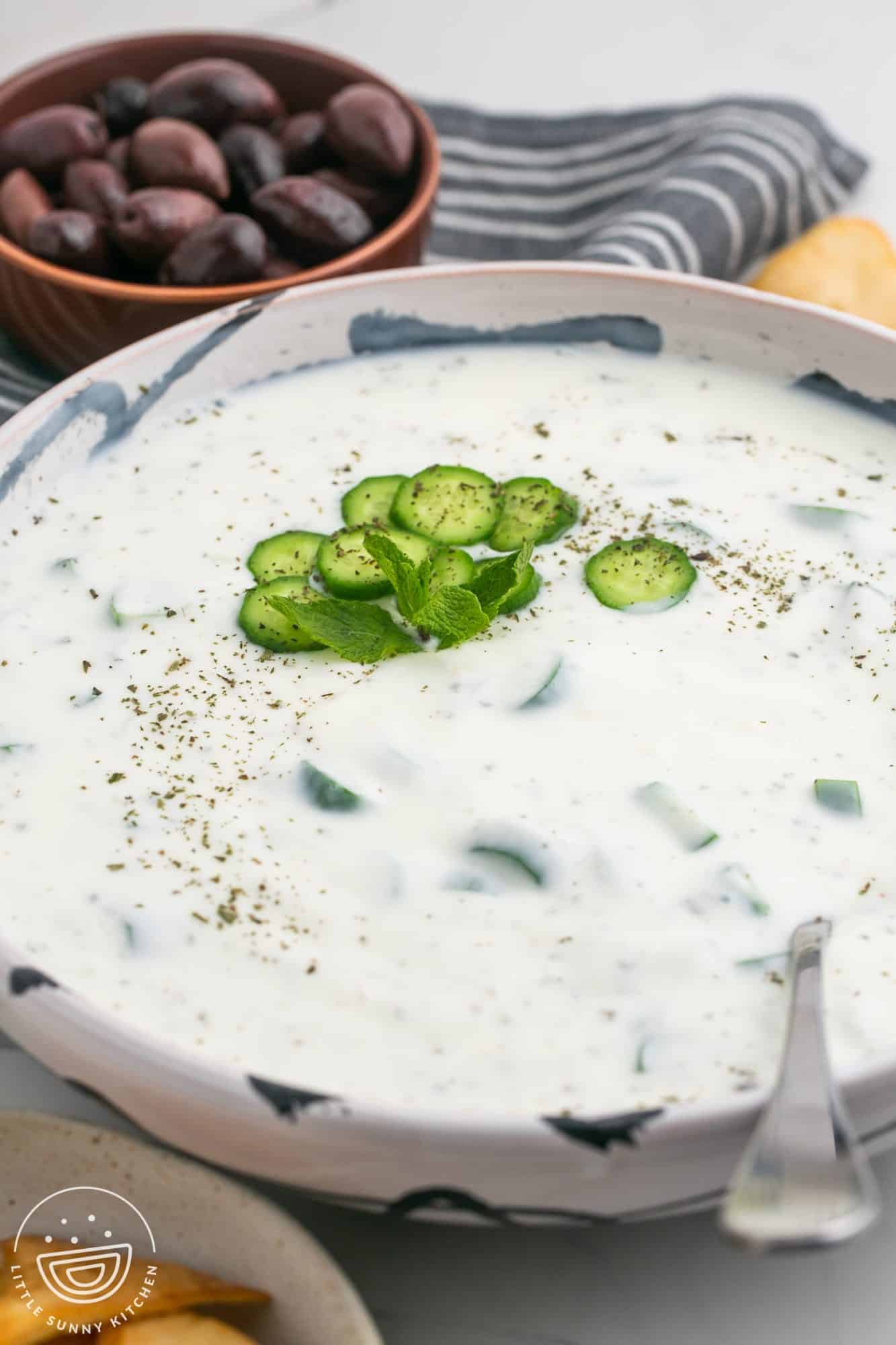 Cucumber yogurt sauce in a large bowl, with sliced of cucumber and dried mint, and a serving spoon. Kalamata olives in the background.