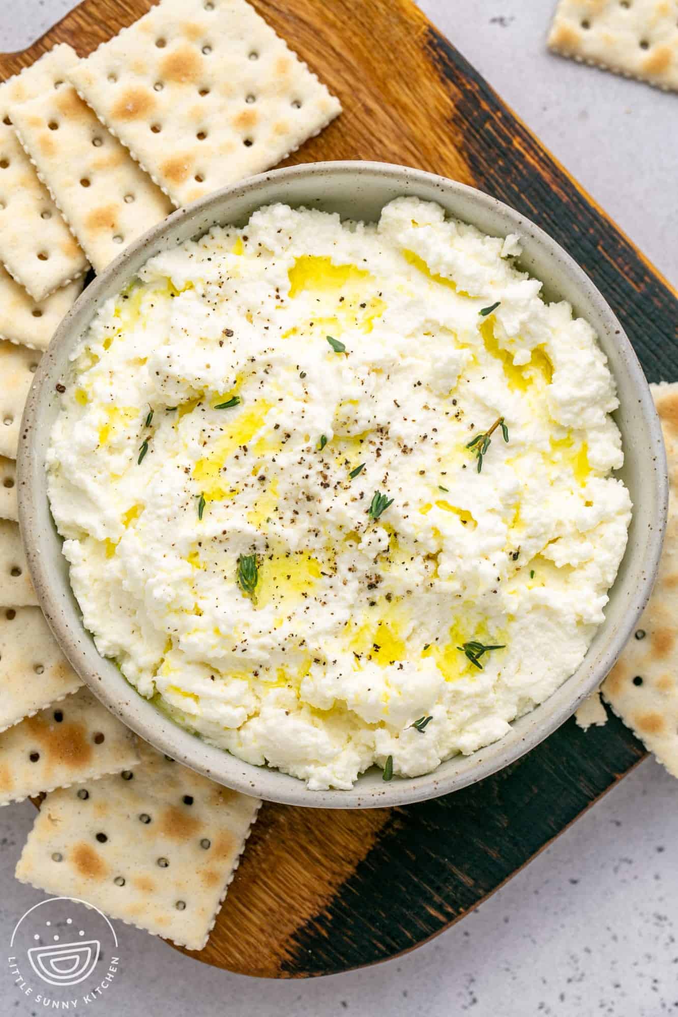 a gray bowl filled with ricotta cheese that has been drizzled with olive oil and sprinkled with pepper and fresh herbs. The bowl is on a wooden board, with saltine crackers around it.