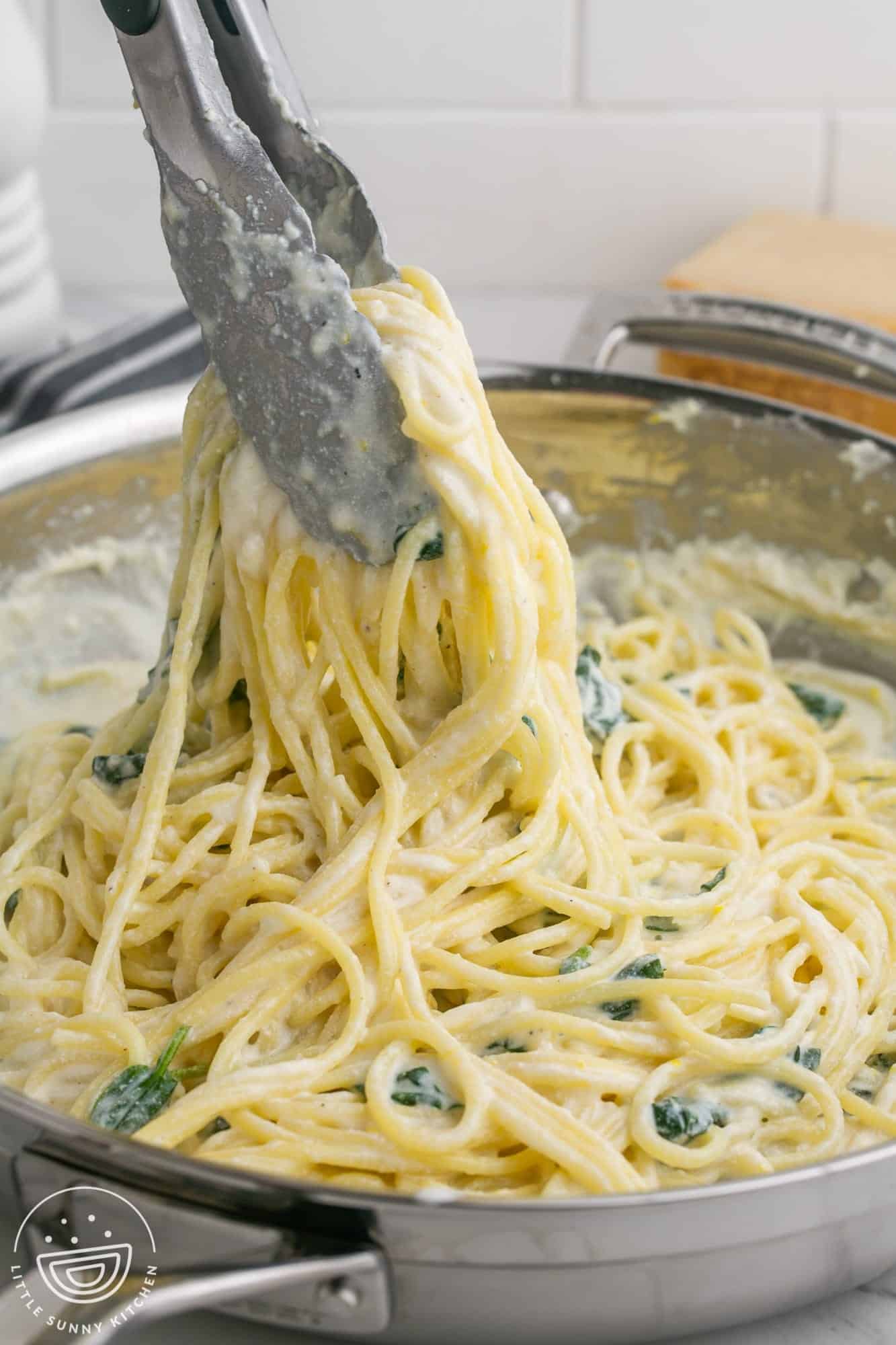 tongs holding up spaghetti with lemon ricotta sauce in a stainless steel skillet. 