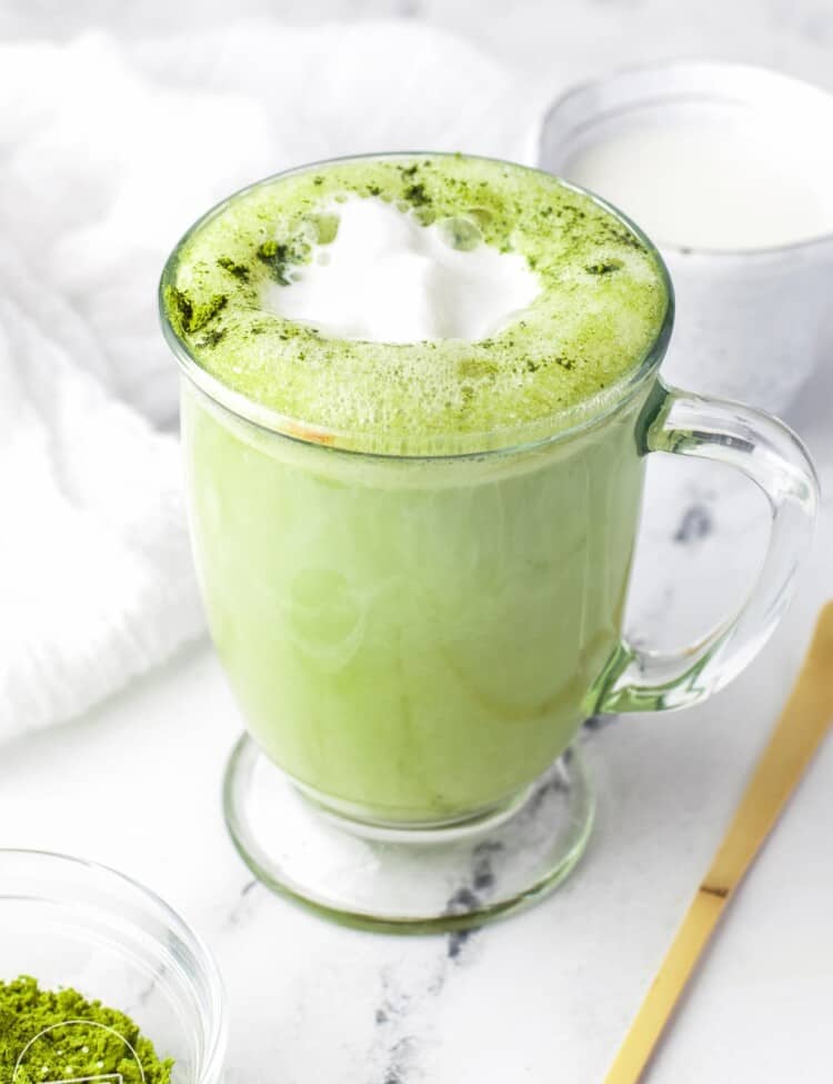 Matcha latte served in a glass mug, and matcha powder in small bowl on the side