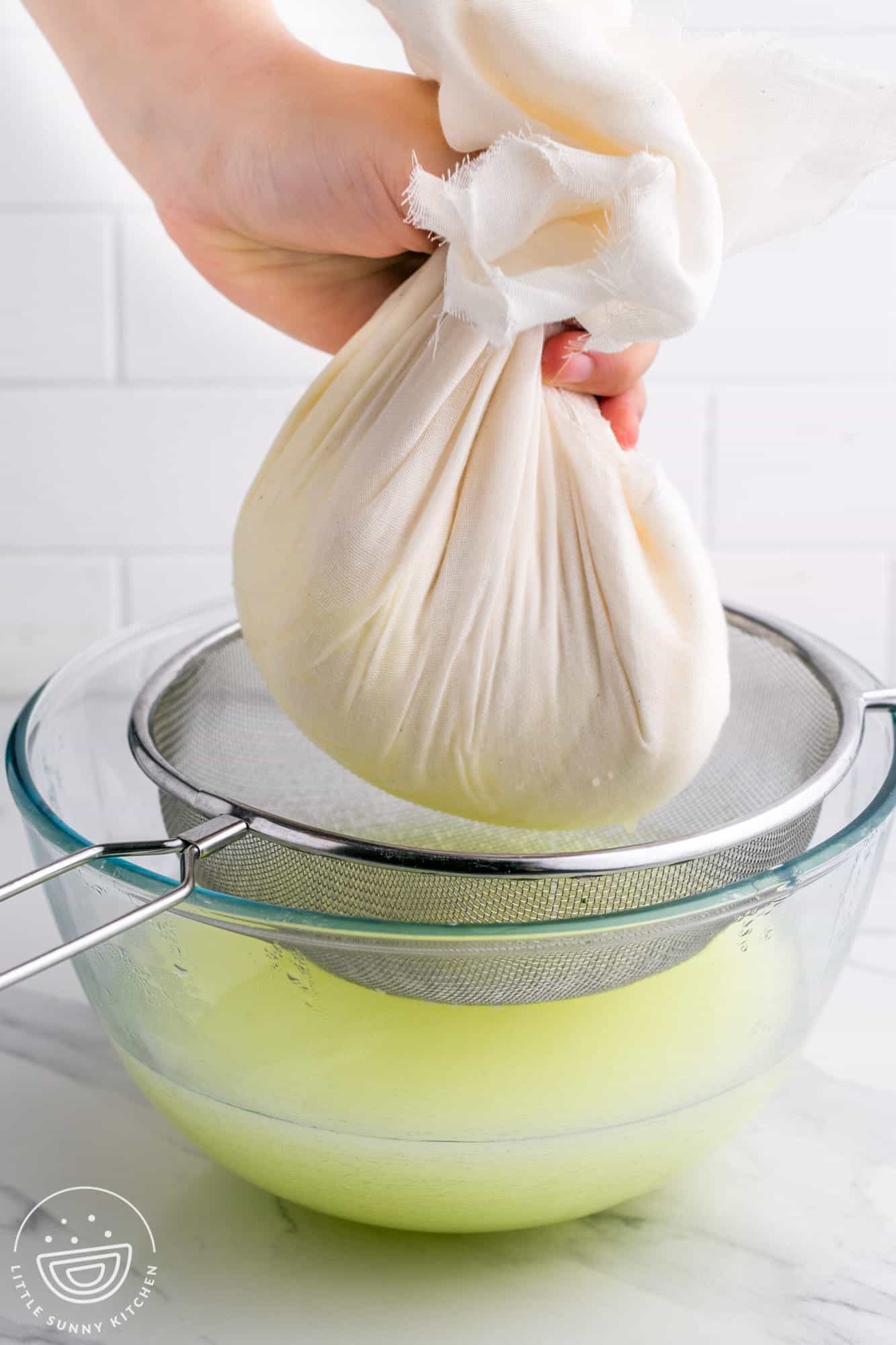 a hand is holding cheese in a cheesecloth over a metal sieve that is over a clear glass bowl. The bowl i filled with whey.
