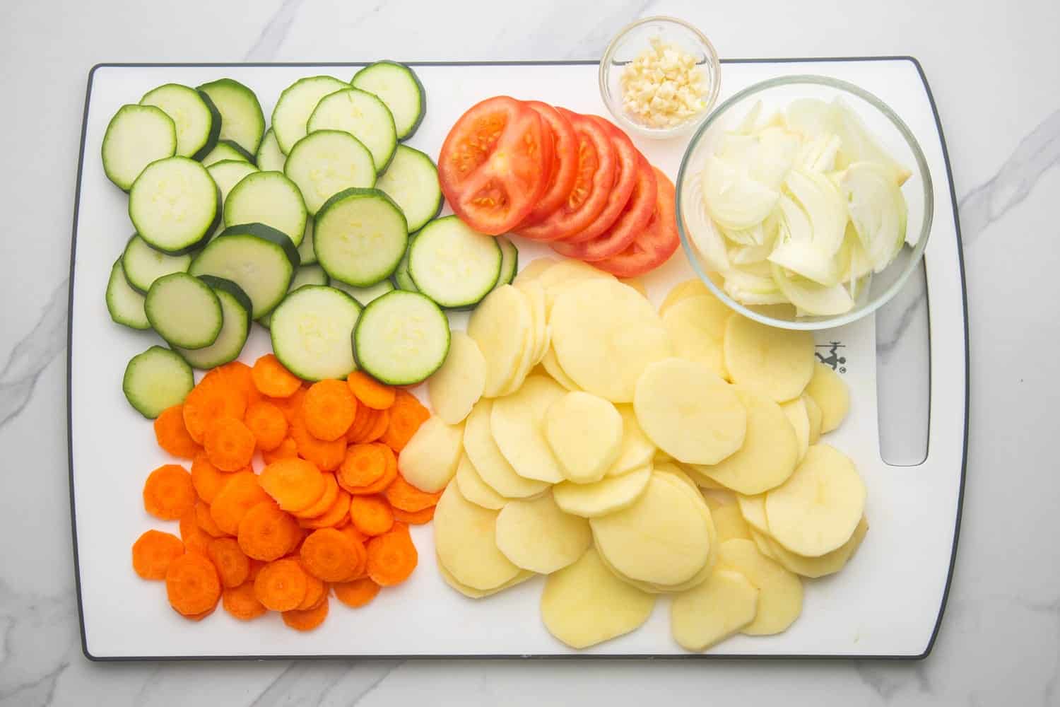 a cutting board holding thinly sliced potatoes, tomatoes, carrots, and zucchini.