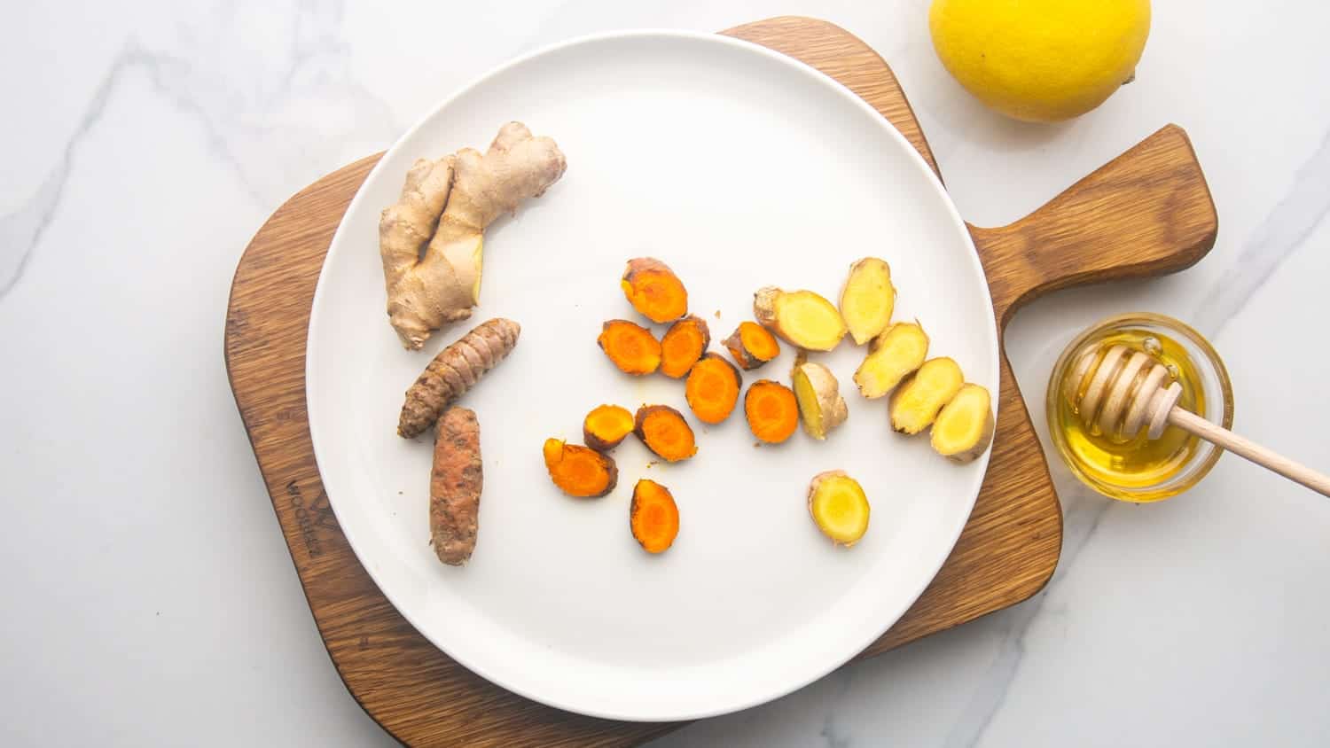 Sliced ginger and turmeric root on a white plate, and honey and lemon on the side