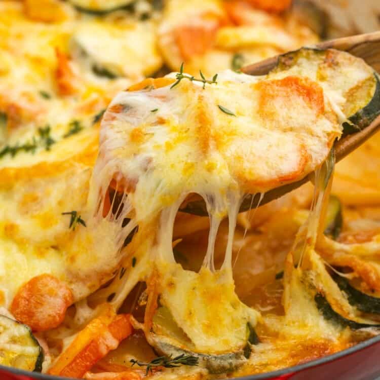 cheesy vegetable potato bake in a round baking dish, a wooden spoon is serving it, showing strings of melty cheese.