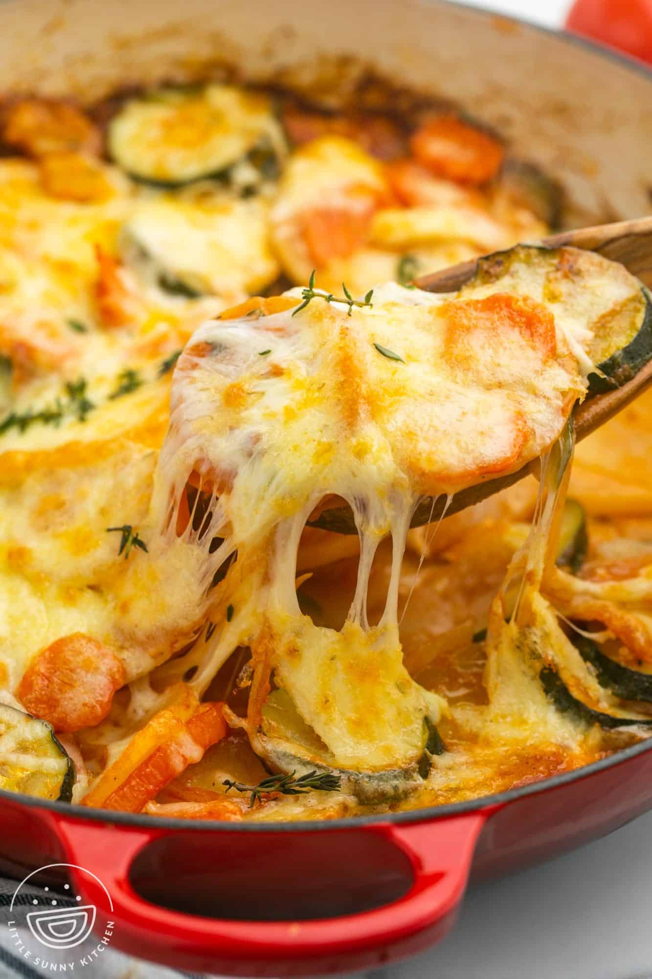 cheesy vegetable potato bake in a round baking dish, a wooden spoon is serving it, showing strings of melty cheese.
