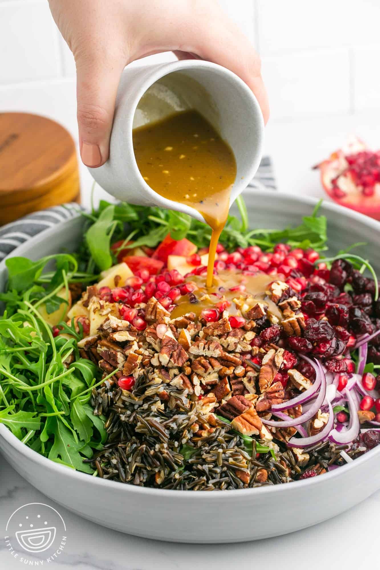 a hand pouring salad dressing over a bowl of arugula (rocket), wild rice and other salad toppings.