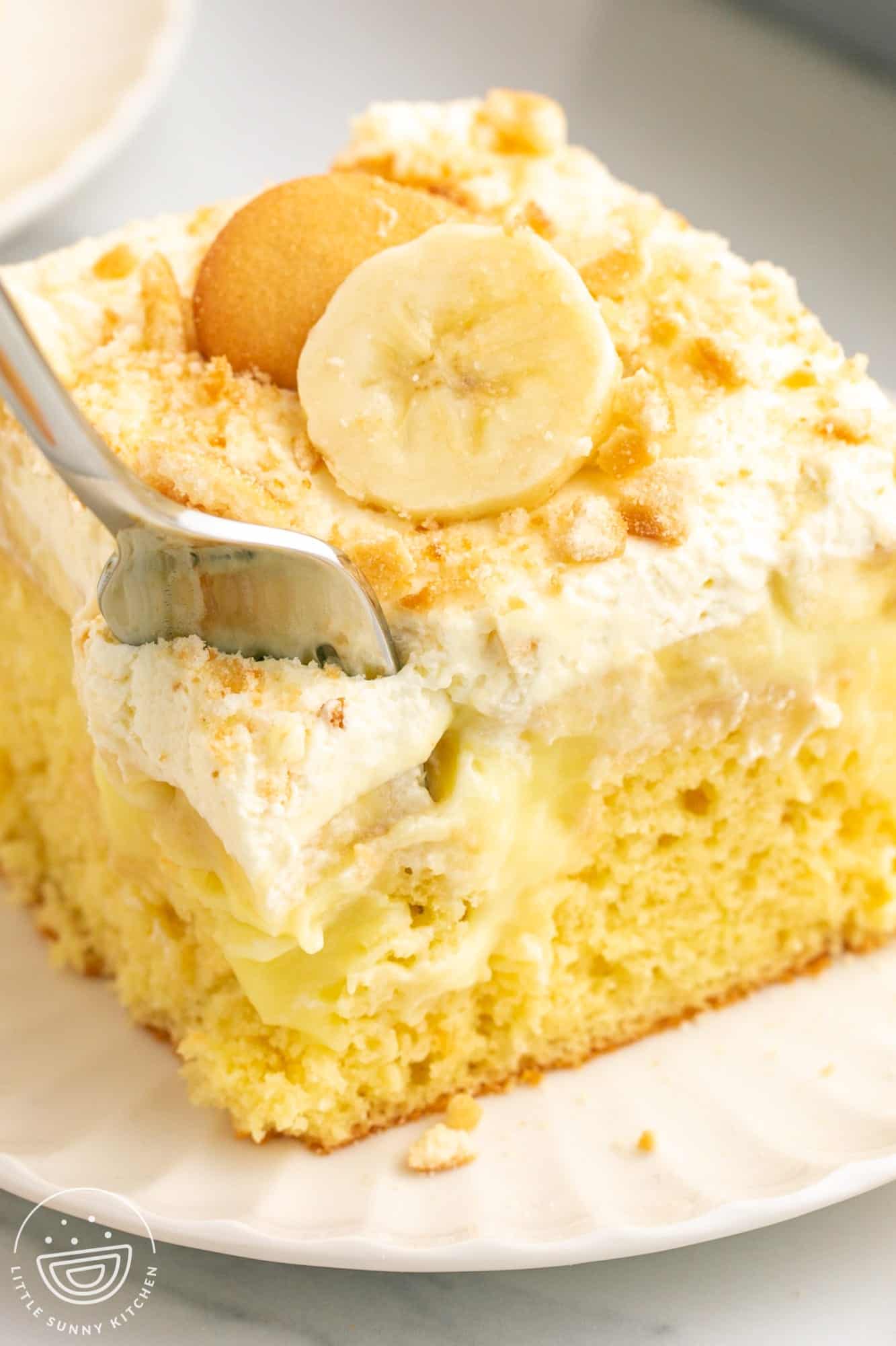 a square slice of banana poke cake on a plate. A fork is taking a bite from the front left corner.