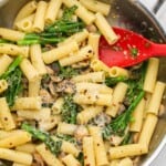 a stainless steel skillet holding rigatoni pasta with broccolini, mushrooms, and parmesan cheese. A red spoon is in the pan as well.