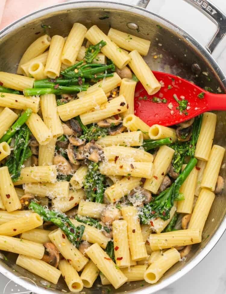 a stainless steel skillet holding rigatoni pasta with broccolini, mushrooms, and parmesan cheese. A red spoon is in the pan as well.