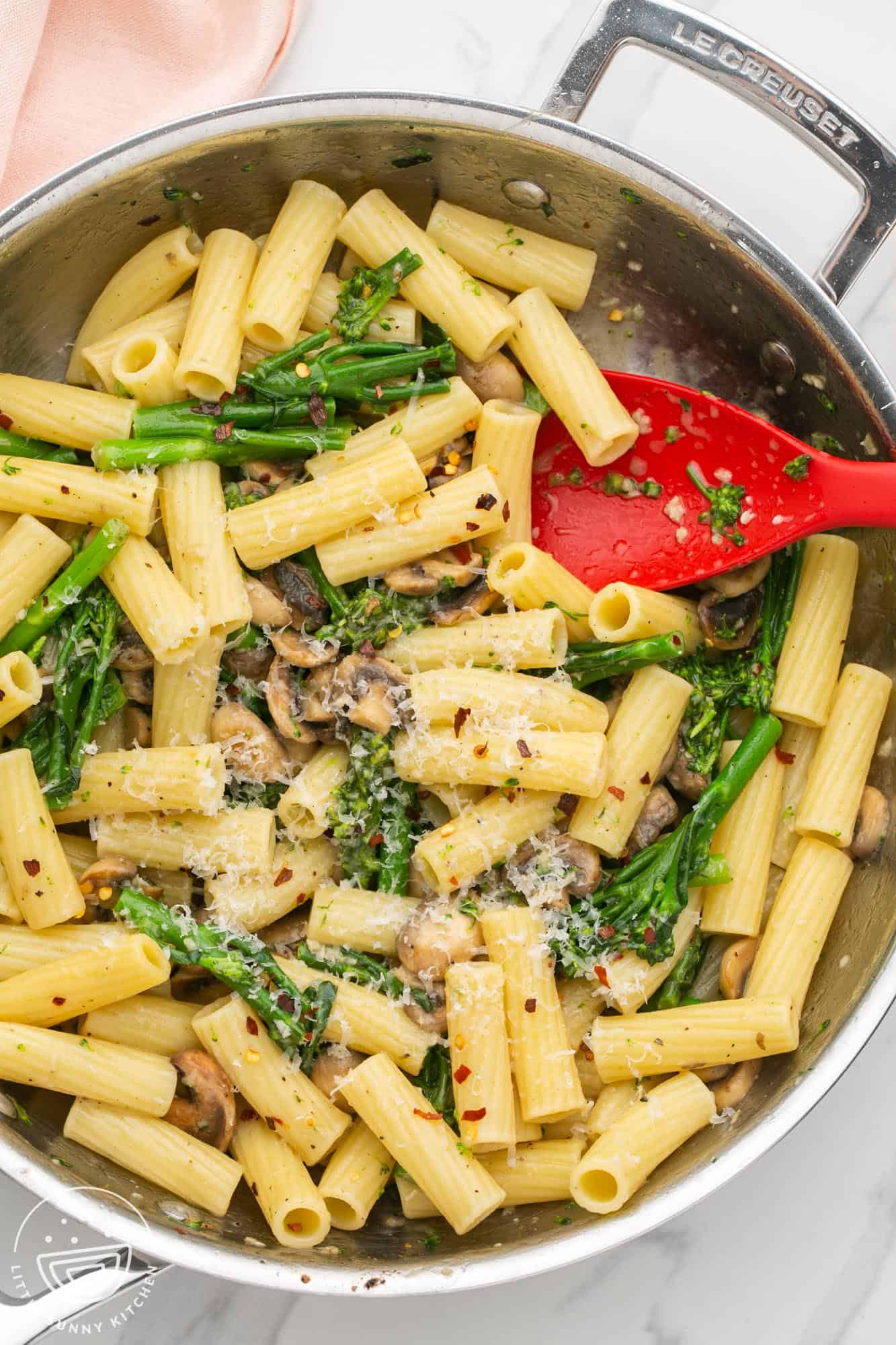 a stainless steel skillet holding rigatoni pasta with broccolini, mushrooms, and parmesan cheese. A red spoon is in the pan as well. 