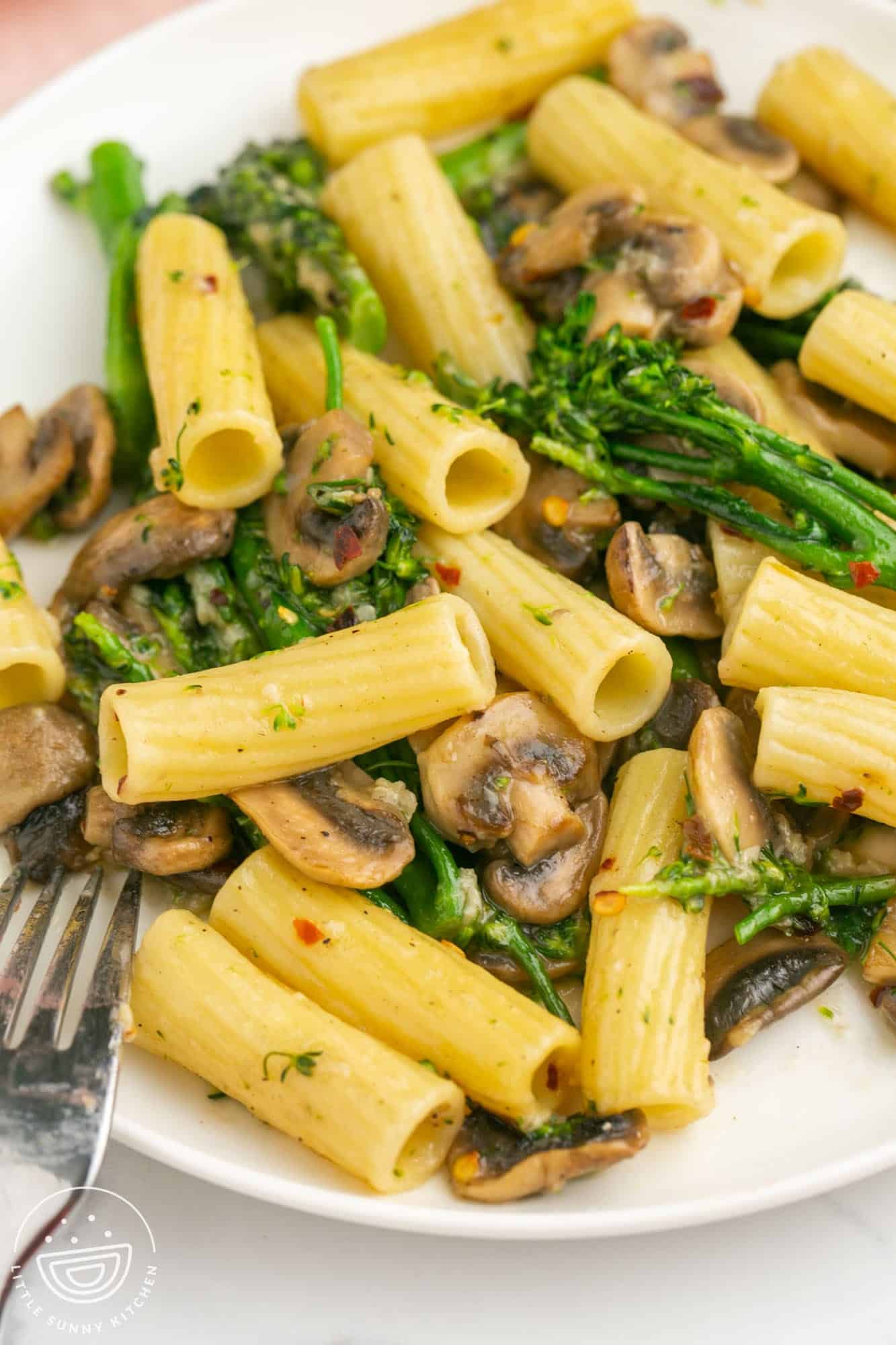 a white dinner plate of tube pasta with mushrooms and broccolini.
