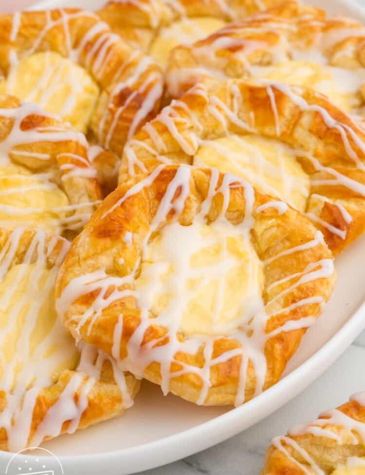 7 cheese danish on an oval platter. They are cheese filled and drizzled with icing.