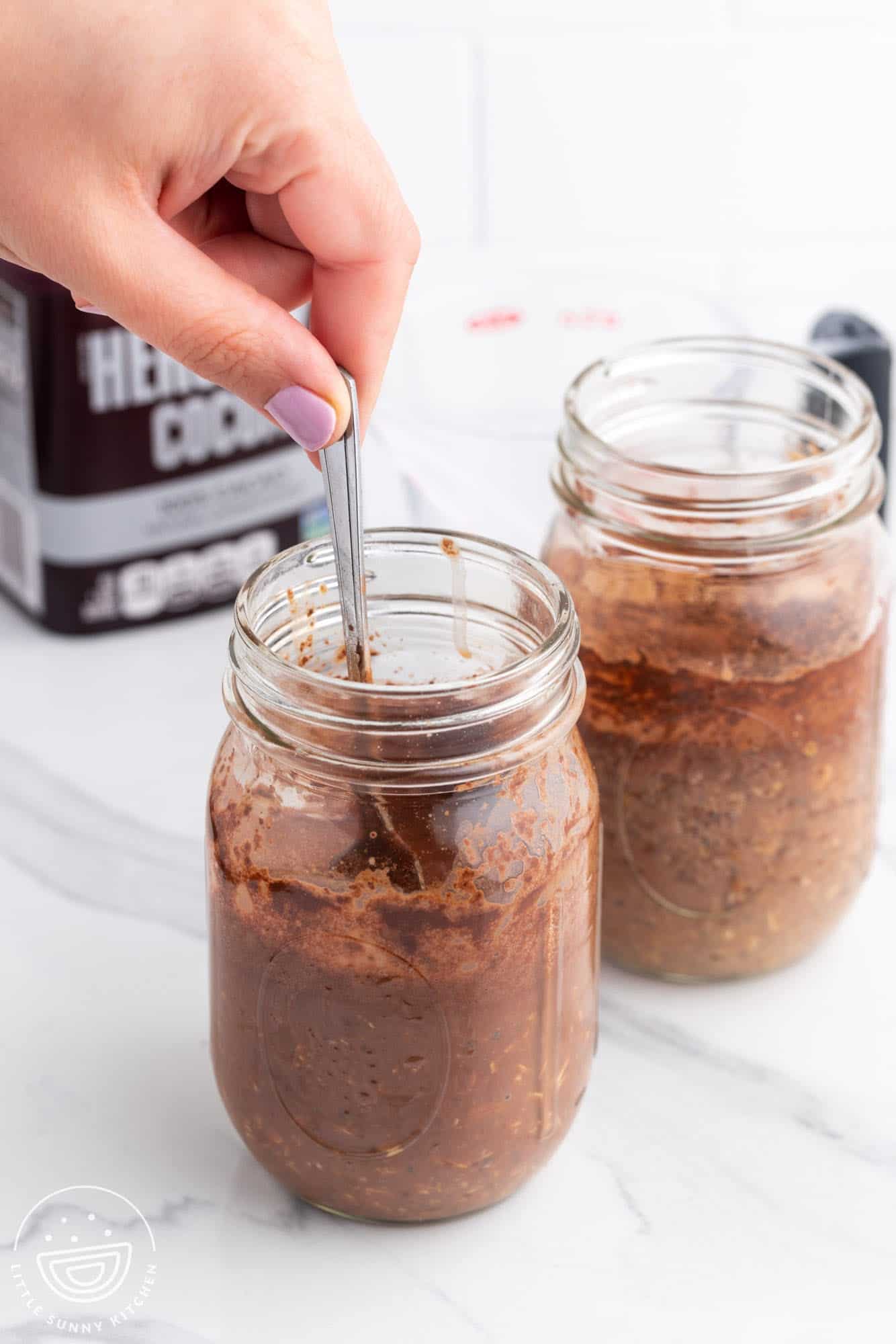 two jars of chocolate overnight oats. a hand is stirring one of them.