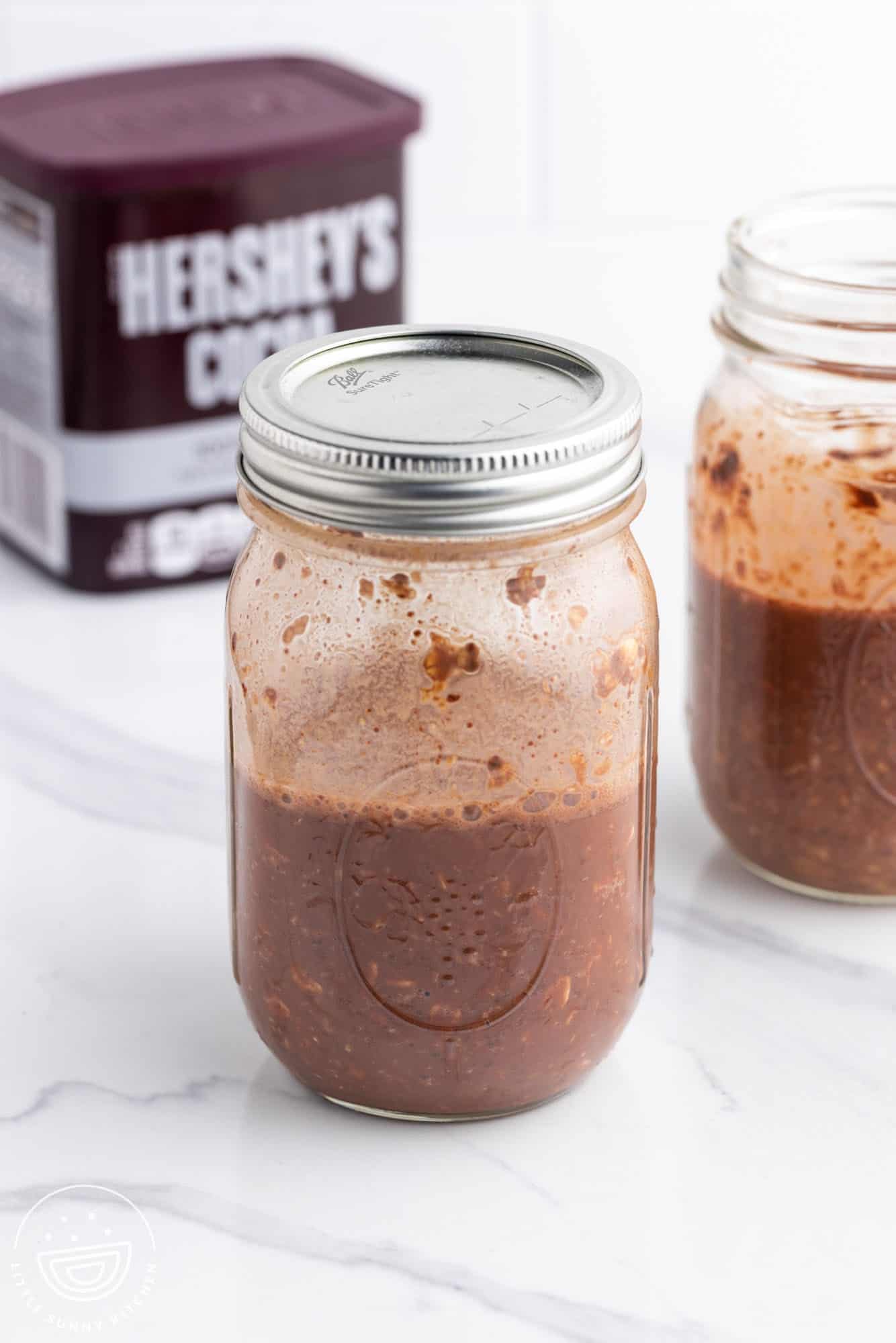 two mason jars of chocolate overnight oats in front of a container of Hershey's cocoa.