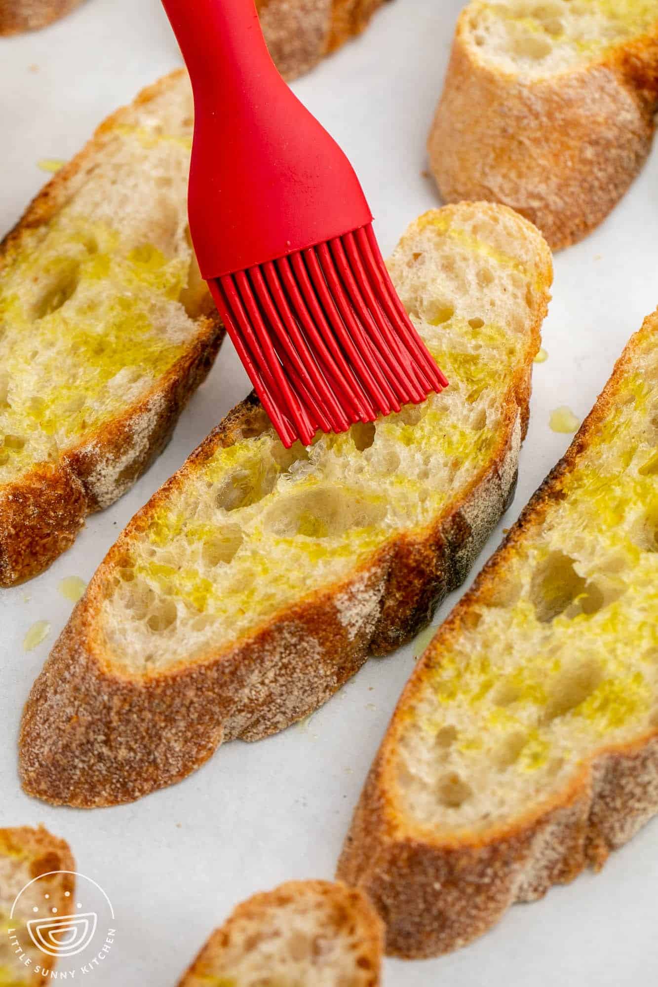 slices of bread on a tray. A red silicone brush is adding olive oil to them.