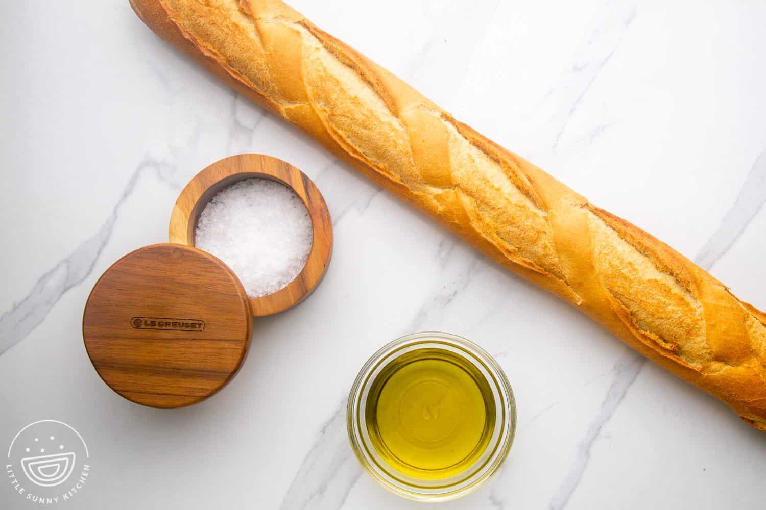 a long baguette next to a wooden tub of salt and a small bowl of olive oil.