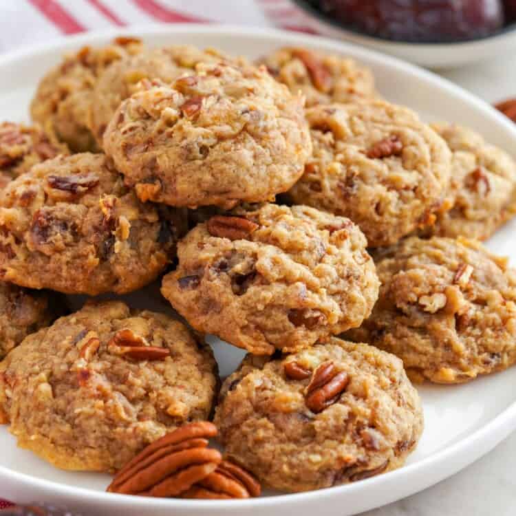 Date cookies with pecans on a white plate. A plate of dates is in the background.