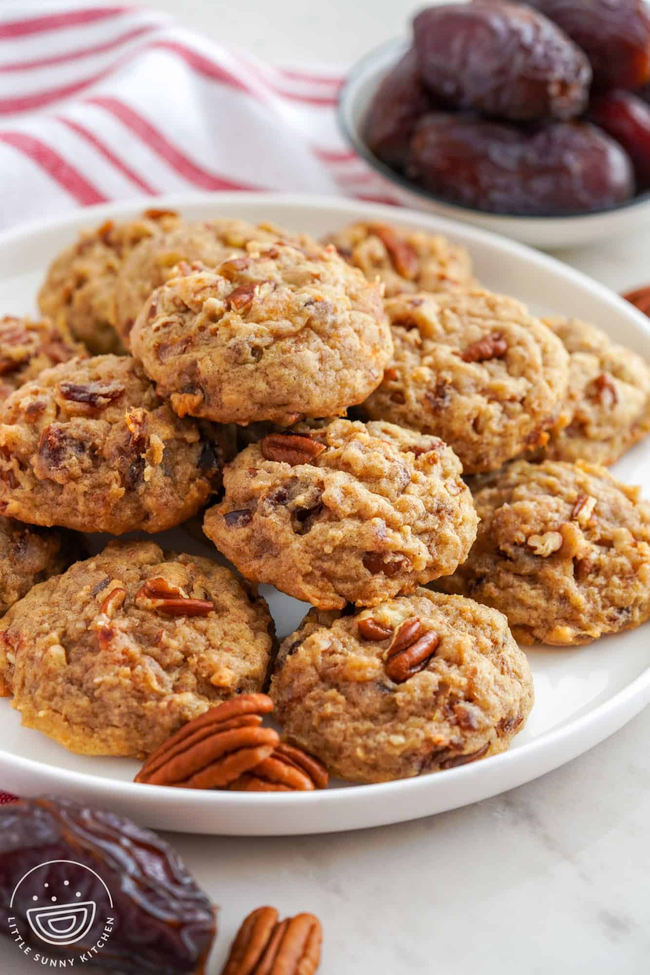 Date cookies with pecans on a white plate. A plate of dates is in the background.