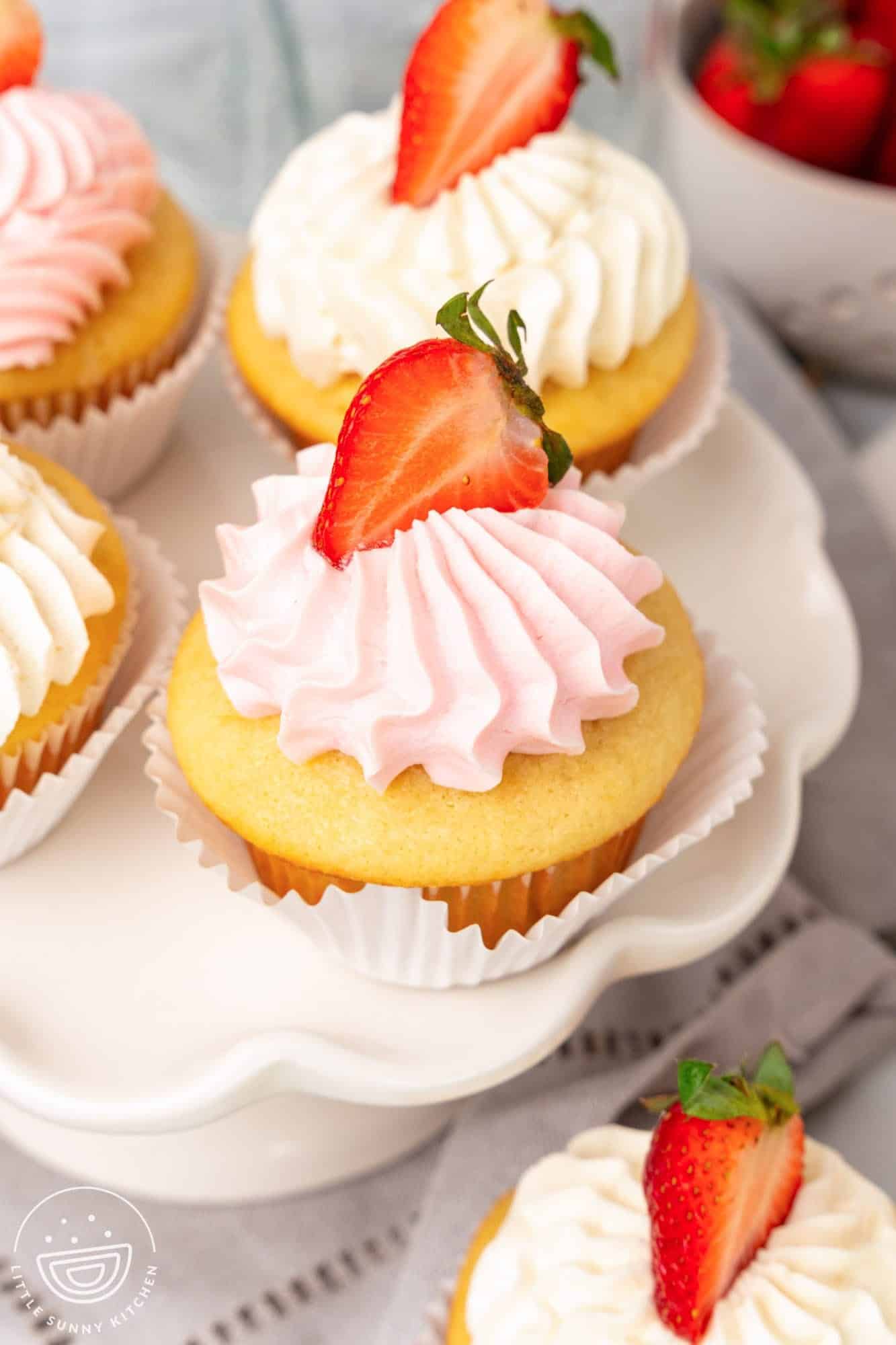 Vanilla cupcakes with pink fluffy frosting, each topped with a half strawberry. 4 Cupcakes are arranged on a small pedestal.