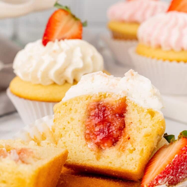 a cupcake cut in half to show strawberry jam in the center. In the background are more cupcakes, decorated with whipped frosting and fresh berries.