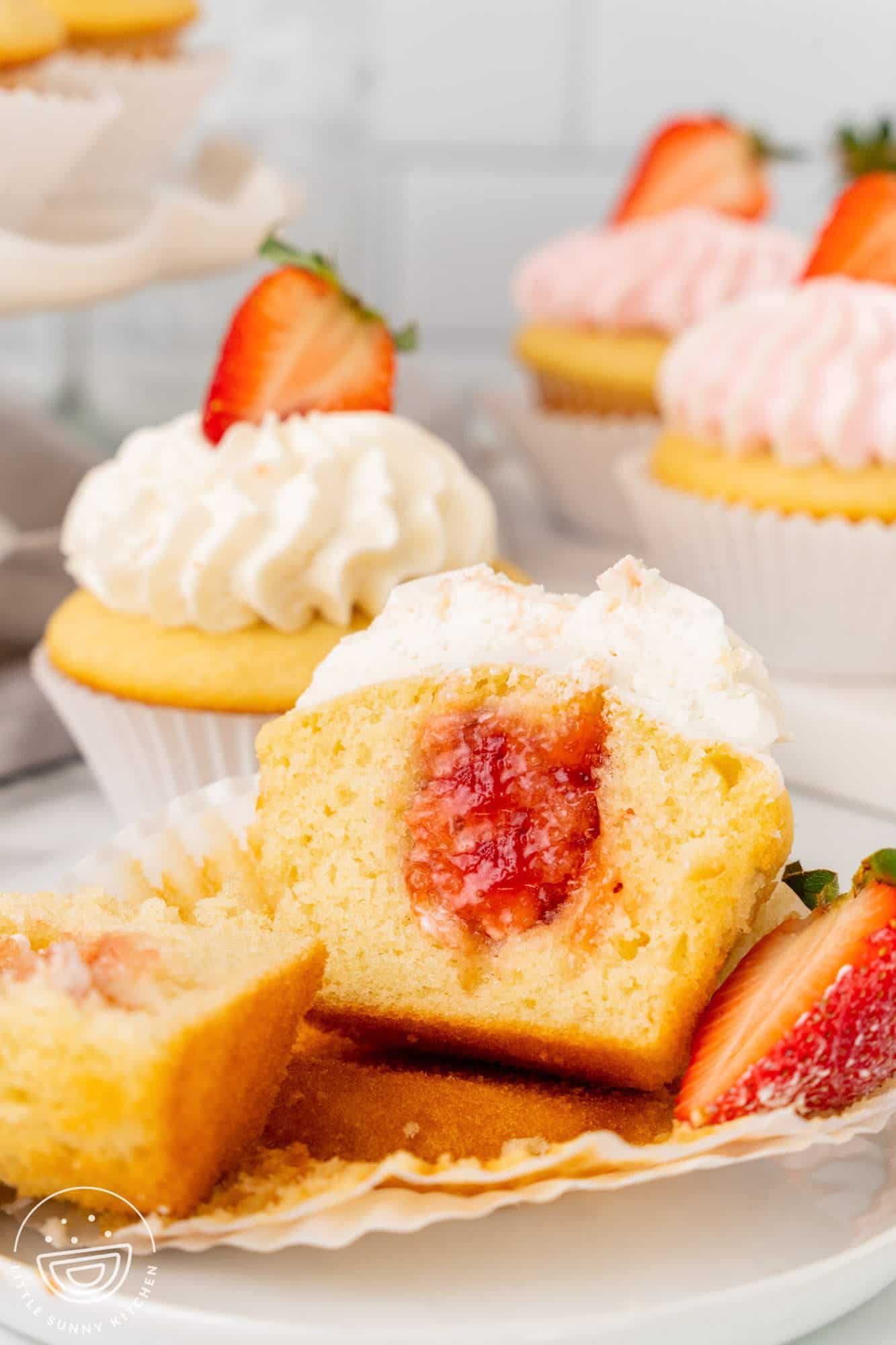 a cupcake cut in half to show strawberry jam in the center. In the background are more cupcakes, decorated with whipped frosting and fresh berries.