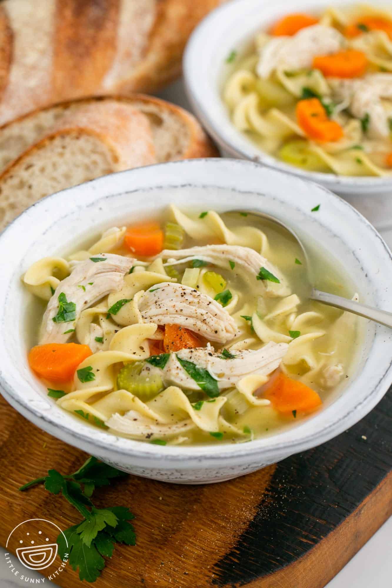a bowl of chicken noodle soup with large pieces of shredded chicken, carrots, celery, and egg noodles. there's a spoon in the bowl and a loaf of bread in the background