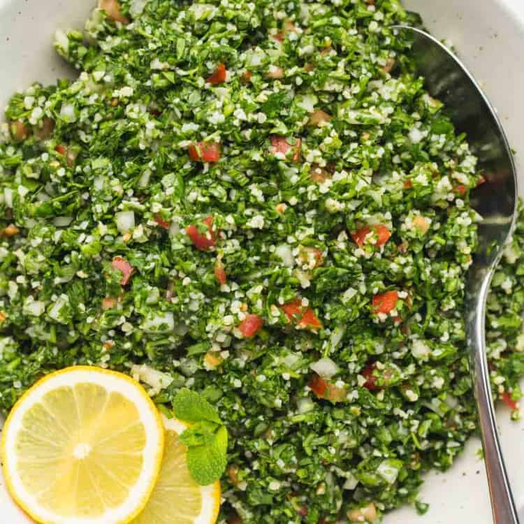 Tabbouleh salad in a white serving bowl with a metal serving spoon and sliced lemons on the side.