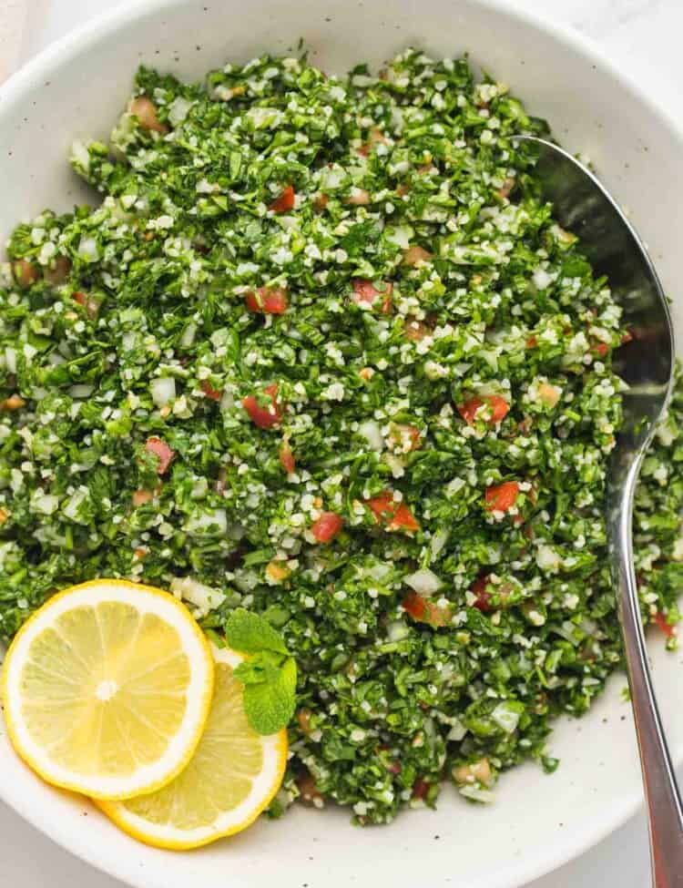 Tabbouleh salad in a white serving bowl with a metal serving spoon and sliced lemons on the side.