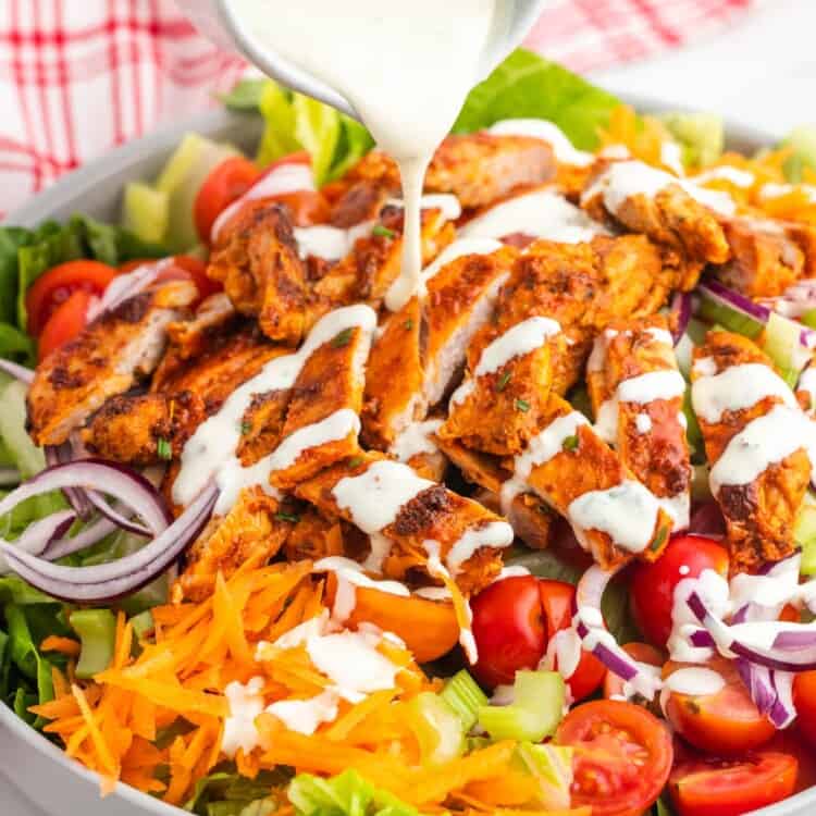 Ranch dressing pouring over a large bowl of salad topped with spicy chicken, shredded carrots, and tomatoes.