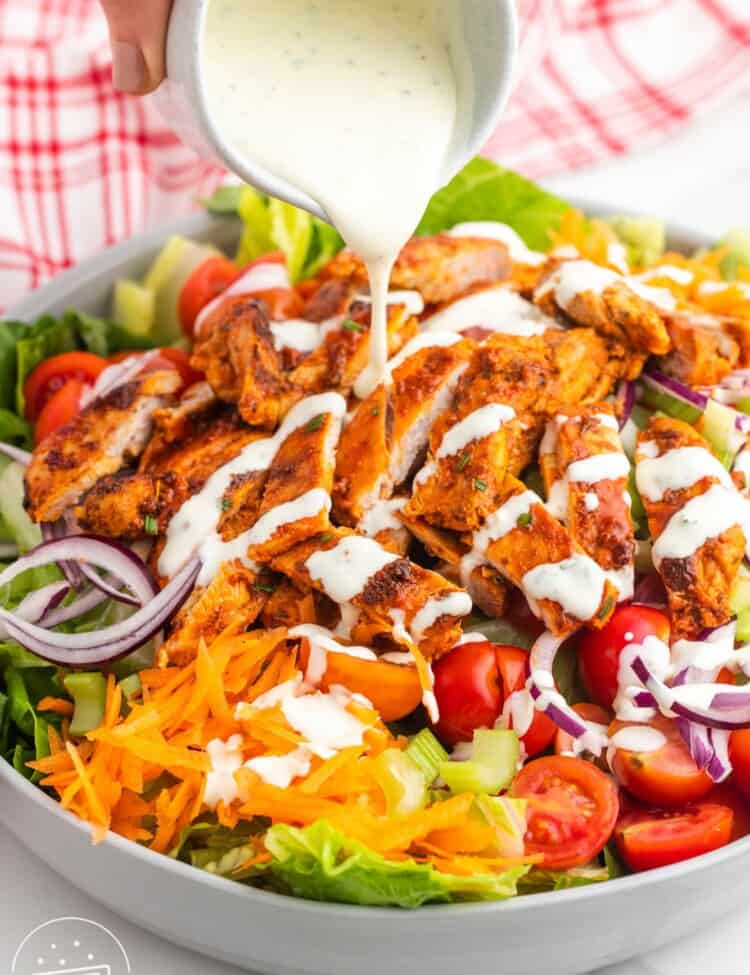 Ranch dressing pouring over a large bowl of salad topped with spicy chicken, shredded carrots, and tomatoes.