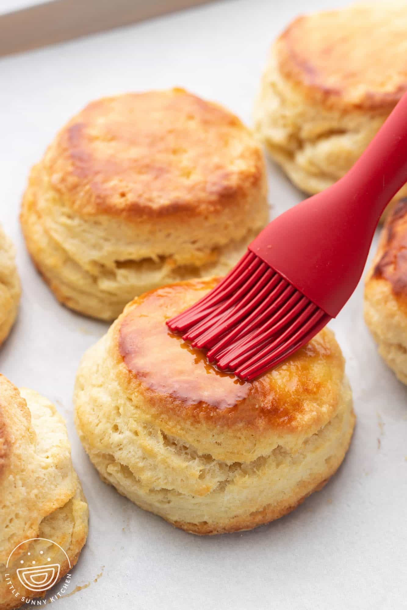 a red silicone brush adding melted butter to the top of a tray of baked buttermilk biscuits.