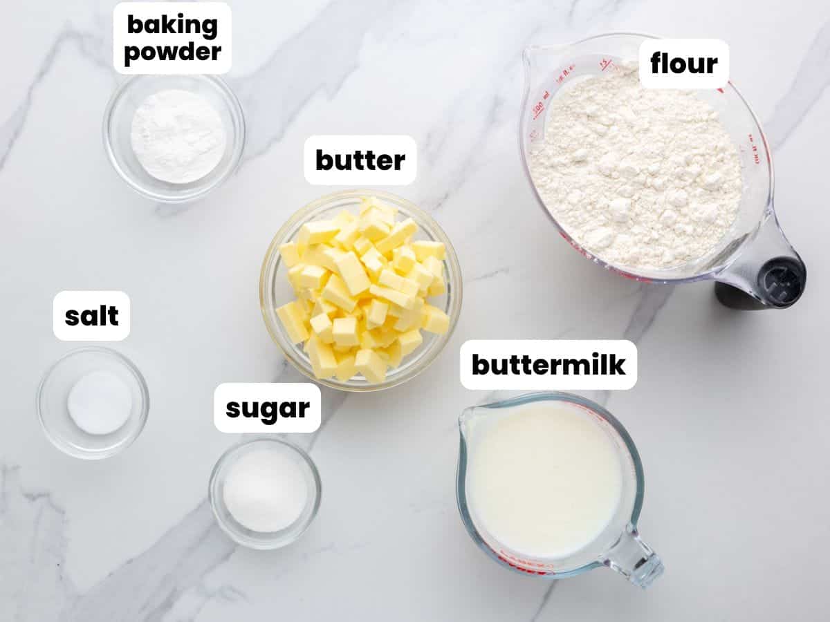 the 6 ingredients needed to make buttermilk biscuits, in separate bowls, arranged on a counter. 