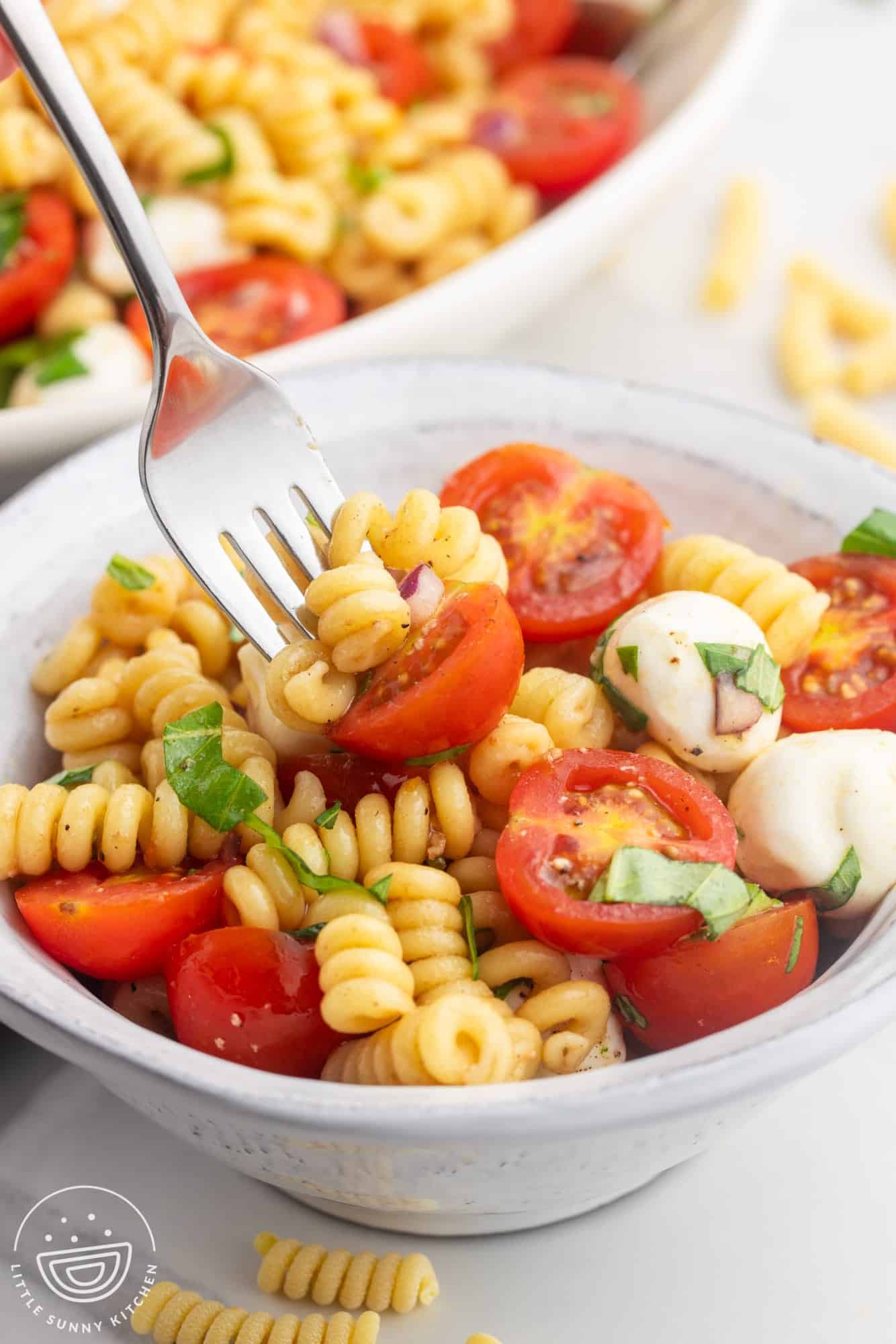 a small white bowl holding a portion of caprese pasta salad. Someone is eating the salad with a fork. 