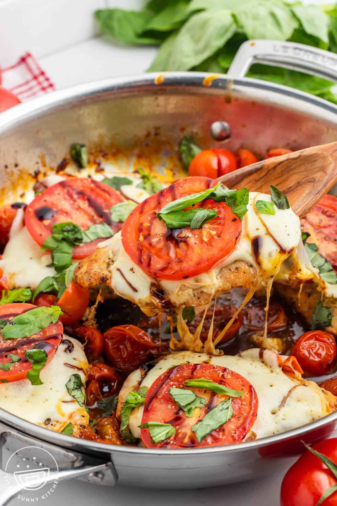 a stainless steel skillet of chicken cutlets topped with caprese tomatoes, cheese, basil. a spoon is lifting one chicken cutlet up out of the pan.