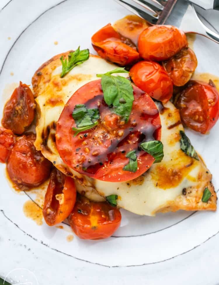 a white plate with a serving of caprese chicken with cherry tomatoes on it.